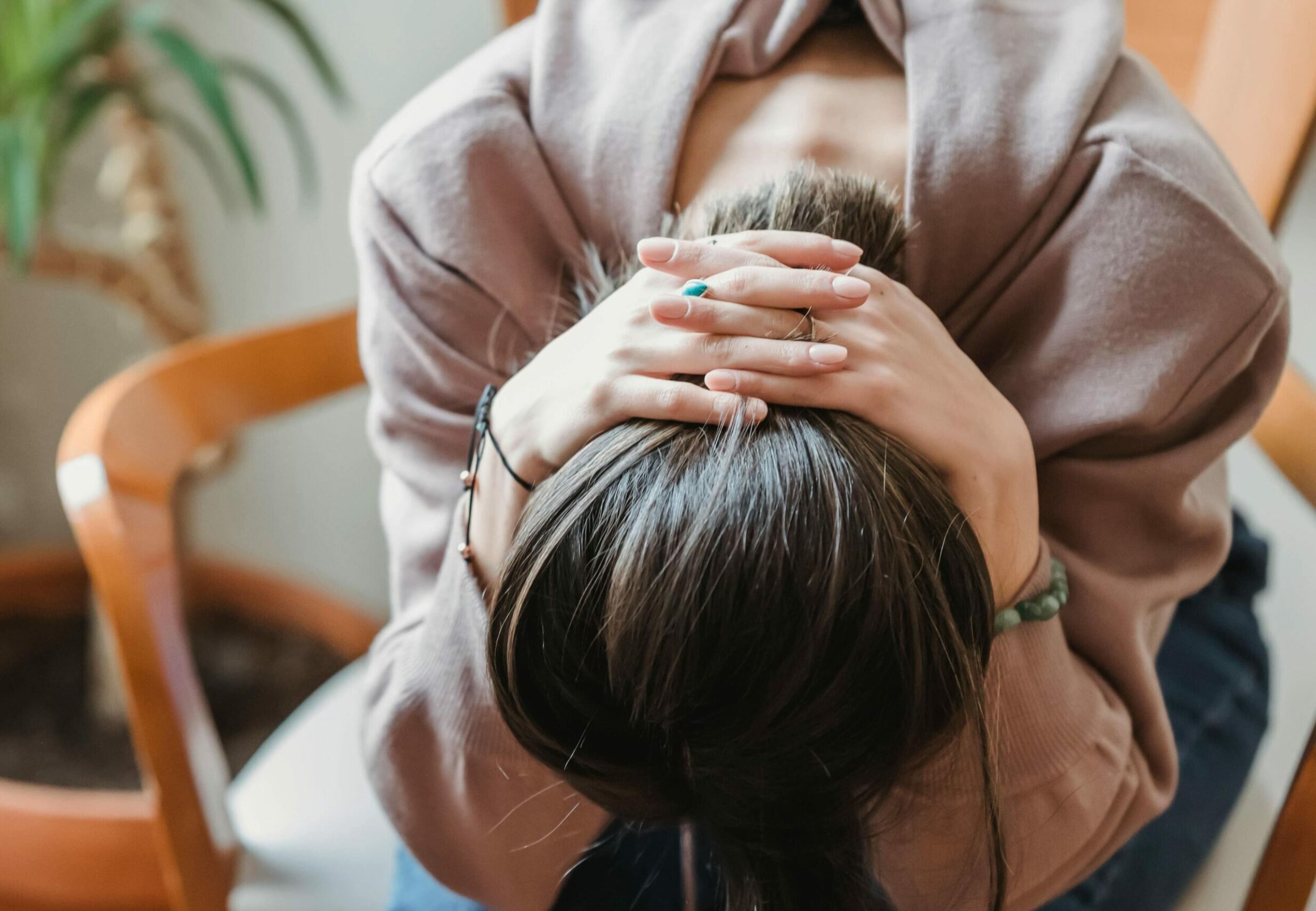Une personne assise sur une chaise en bois est penchée en avant avec la tête entre les mains. Les mains sont jointes derrière la nuque, les coudes pointant vers l'extérieur. La personne porte un sweat-shirt rose clair et un jean. Ses longs cheveux bruns sont attachés en queue de cheval. On aperçoit un bracelet bleu clair à son poignet gauche. Un bout de plante verte est visible en haut à gauche de l'image. Le fond est clair et flou.